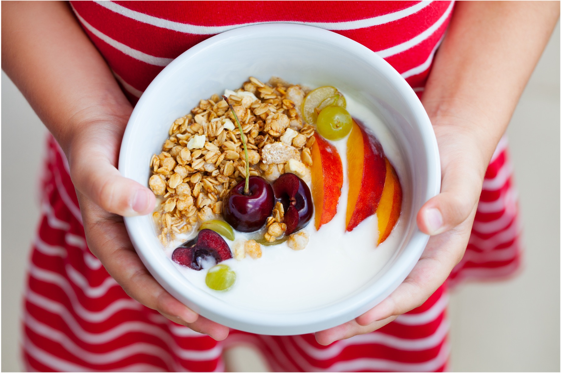 A child in a red and white striped dress holds bowl of granola and fruit