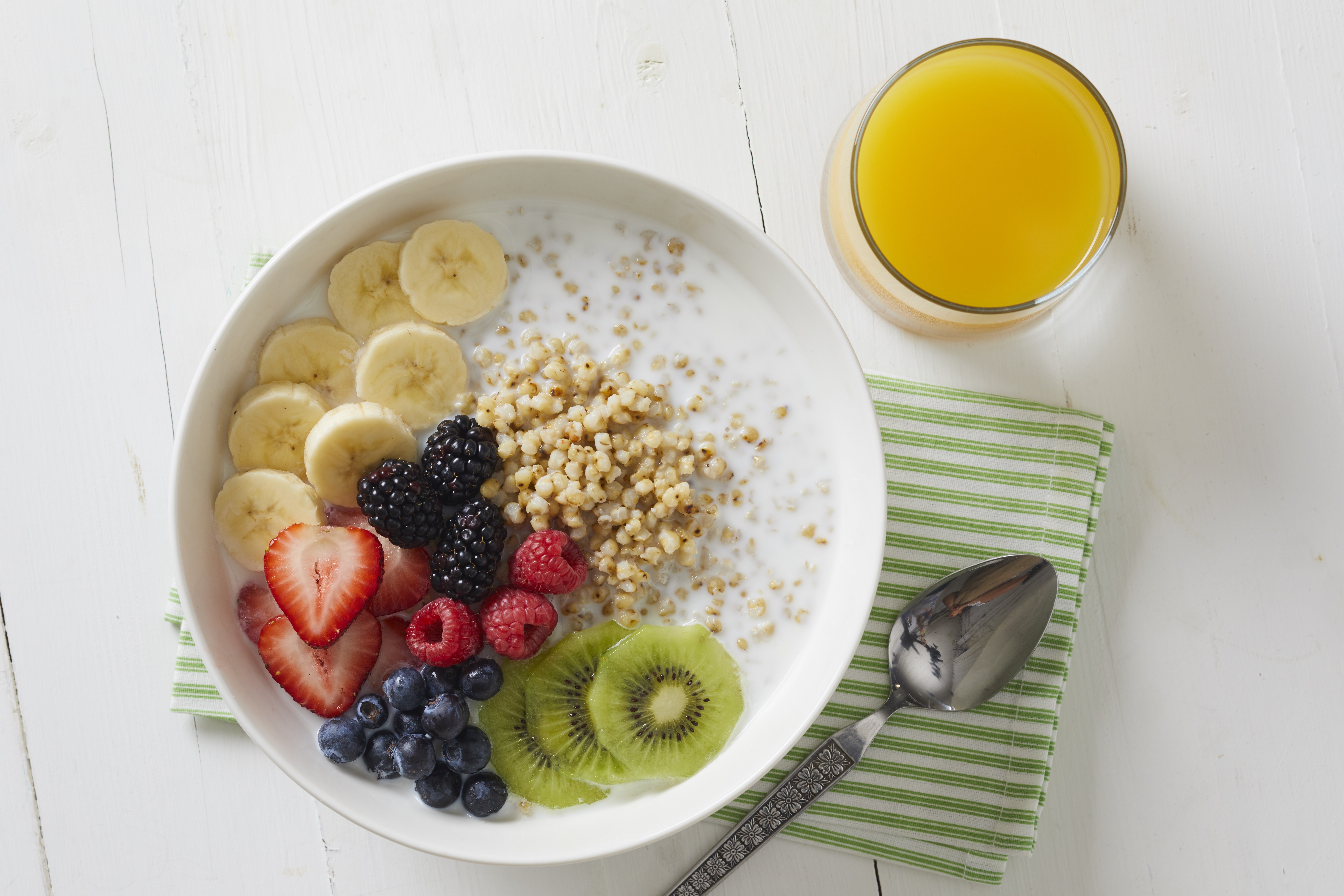 A bowl of cooked sorghum grain with milk, topped with bananas, strawberries, kiwi, and bluberries on a table with orange juice and a spoon on a green napkin