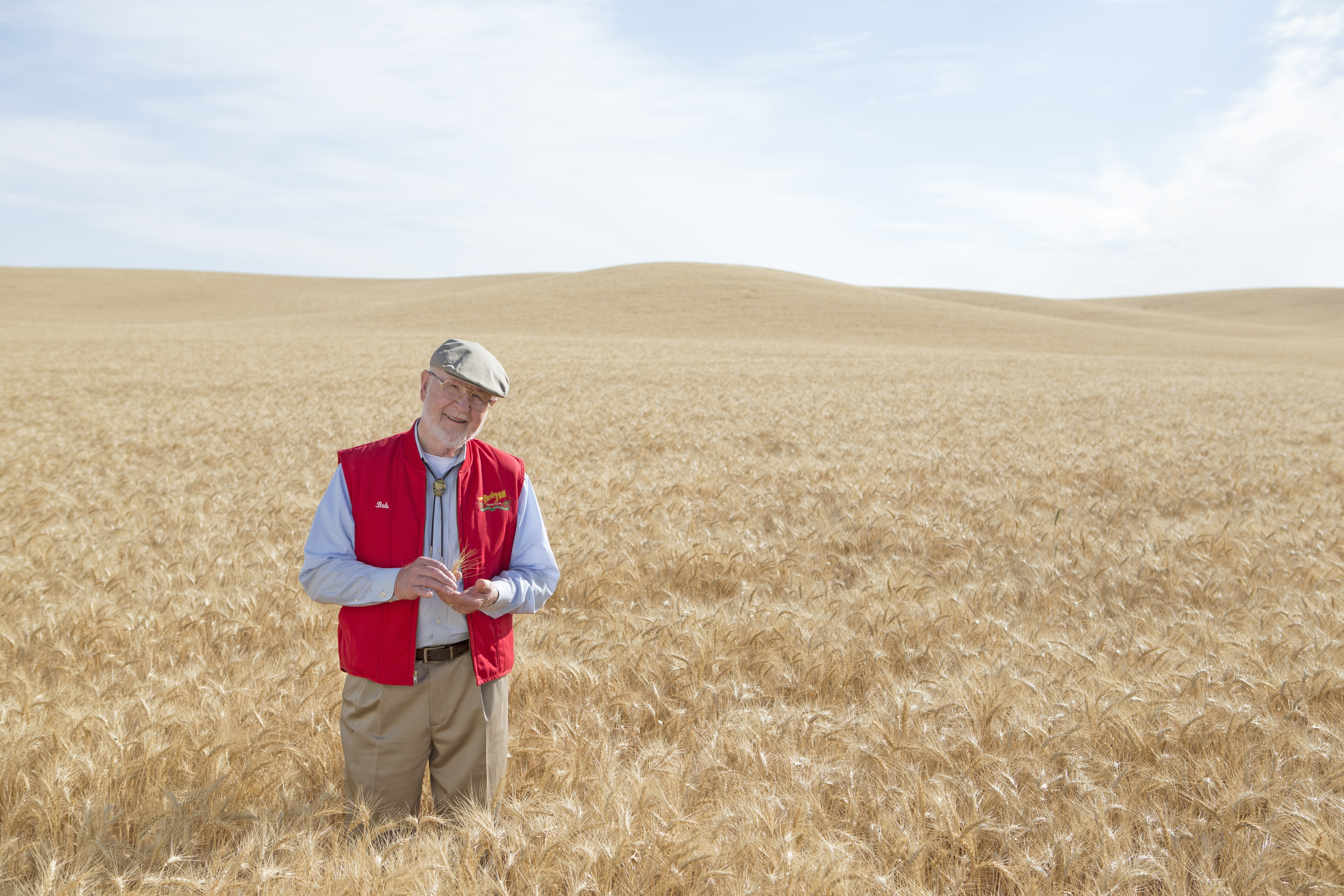 Bob Moore standing in a golden field of grain. The grain fills the foreground and background, and the sky is blue and cloudy.