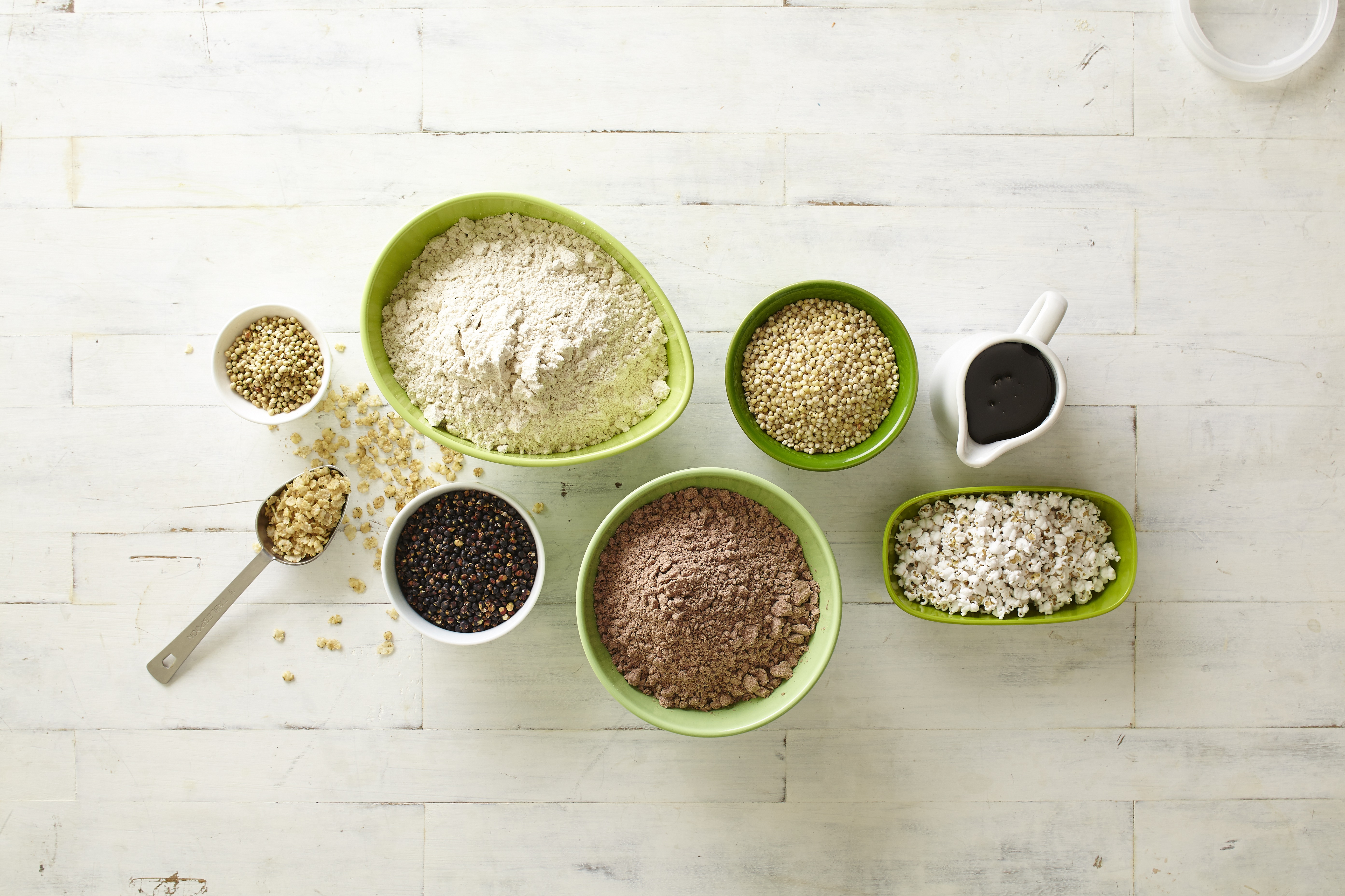 different forms of sorghum - white, black, sorghum flour, sorghum syrup, popped sorghum and flaked sorghum -  in green bowls on a white table